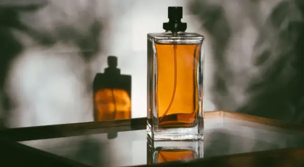 Szklany flakon perfum z bursztynowym płynem i czarną nakrętką, odbijający się w lustrzanej powierzchni.