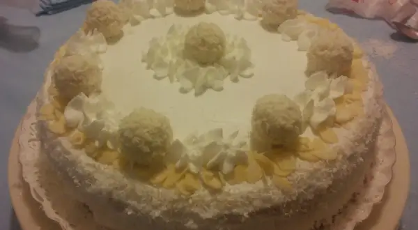 Torta Raffaello ricoperta di cocco, decorata con panna montata e praline, un'esplosione di dolcezza.