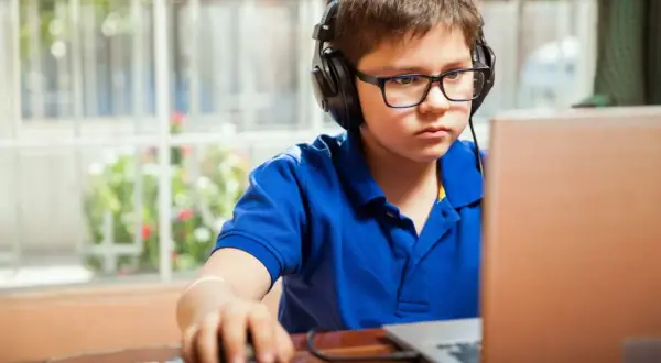 Chłopiec w okularach i słuchawkach skupiony na grze na laptopie. Dziecko przy komputerze z myszką w dłoni.