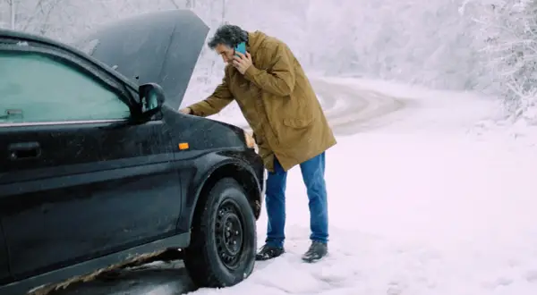 Man med telefon vid en bil med öppen motorhuv. Kallstart bil i snöväder.