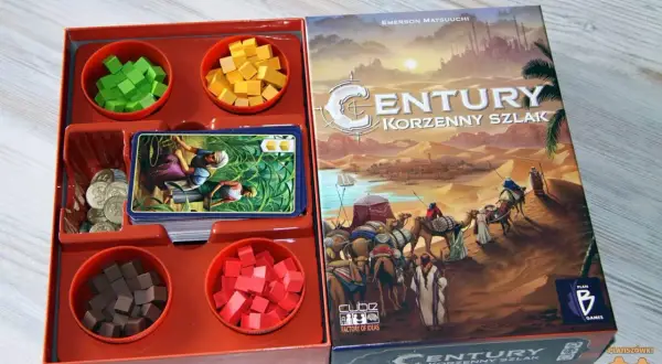Planszówka "Century: Korzenny szlak" z kolorowymi kostkami, kartami i monetami. Karawana wielbłądów przemierza pustynię.