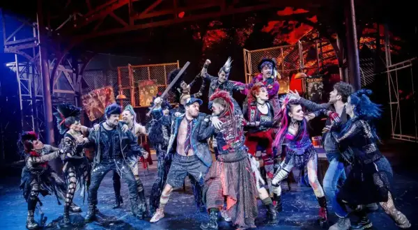 Grupa punkowych artystów na scenie, gotowa do występu. Ich stroje i fryzury krzyczą "we will rock you tłumaczenie" w każdym detalu.