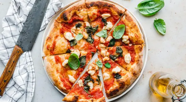 Eine perfekt gebackene vegane Pizza mit Tomaten, veganem Mozzarella und Basilikum, daneben ein Messer und Olivenöl.