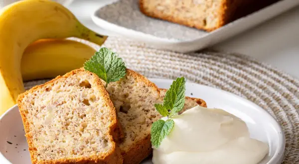 Slices of banana bread with cream and mint leaves, next to whole bananas.