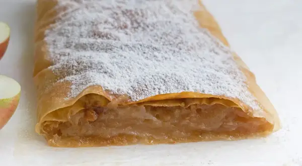 Ein köstlicher Apfelstrudel mit fertigem Strudelteig, bestäubt mit Puderzucker. Daneben liegen frische Apfelstücke.