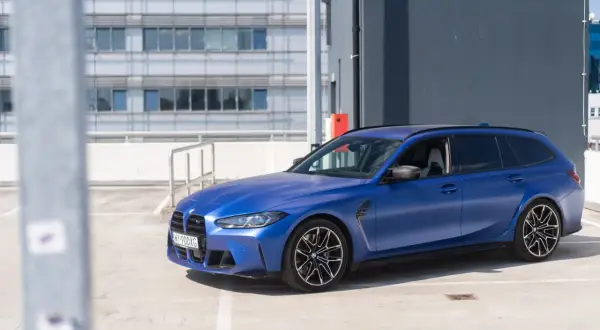 Niebieskie BMW M3 Touring na parkingu. Sportowy kombi z matowym lakierem prezentuje się dynamicznie na tle nowoczesnego budynku.