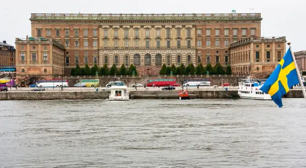 Stor byggnad vid vattnet, troligen kuninkaanlinna tukholma, med bussar och båtar. Svenska flaggan vajar.