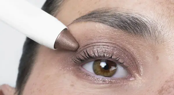 Application d'un fard à paupières bronze sur des yeux marrons. Un bâtonnet blanc dépose la couleur sur la paupière.