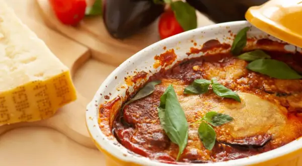 Gebackene Auberginen mit Tomatensoße und Basilikum in einer Auflaufform, daneben Parmesan und frische Auberginen.