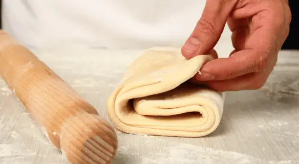 Mani che piegano la pasta sfoglia fatta in casa, con un mattarello in legno accanto, su un piano infarinato.