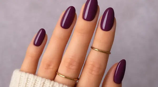 Main d'une femme aux ongles vernis d'un prune profond, une parfaite idée ongle janvier. Deux fines bagues dorées ornent les doigts.