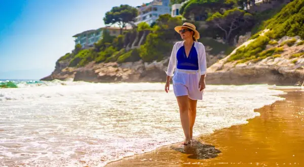 Kobieta w słomkowym kapeluszu spaceruje po plaży. Idealne miejsce na tanie loty na majówkę, by poczuć słońce i fale.