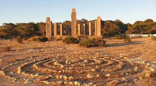 Steine bilden ein Labyrinth im Freien, umgeben von Pinien. Im Hintergrund ragen steinerne Säulen auf, die an Zeit und Raum auf Ibiza erinnern.