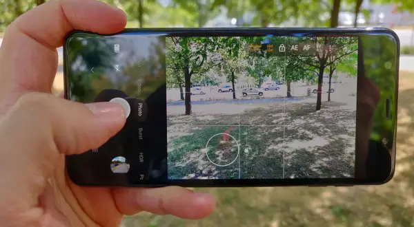 Dłoń trzyma smartfon z otwartą aplikacją do robienia zdjęć. Widok na drzewa i samochody.