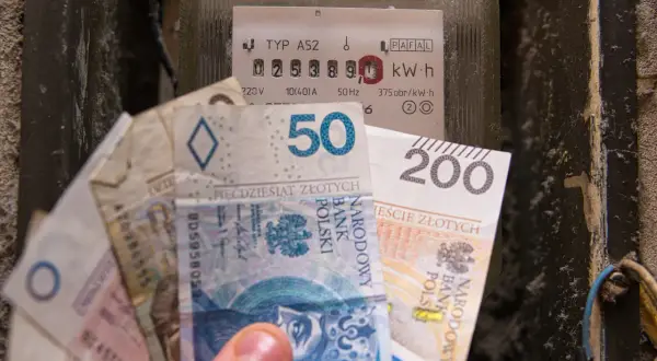 Dłoń trzyma polskie banknoty przy liczniku energii elektrycznej. Zastanawiasz się, po ile jest prąd?