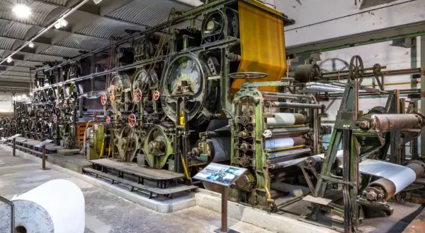 Große, grüne Textilmaschine im lvr industriemuseum, die Stoffbahnen verarbeitet. Eine gelbe Stoffbahn hängt oben, daneben Papierrollen.