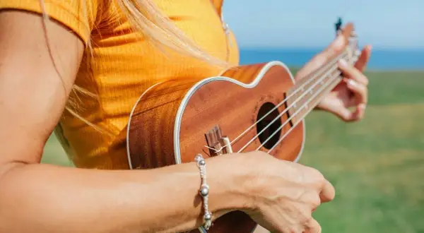 Kobieta w żółtej koszulce gra na ukulele na tle zielonej łąki i morza.
