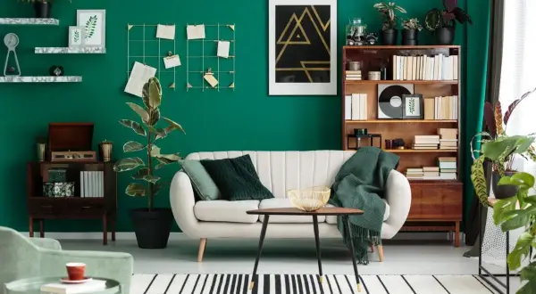 Soggiorno con parete verde smeraldo, divano bianco, libreria in legno e piante che creano diverse **tipi di green**.