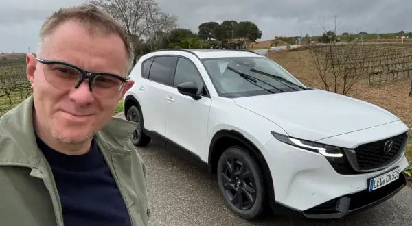 Mężczyzna w okularach pozuje do selfie obok białej Mazdy CX-5 na tle winnicy.