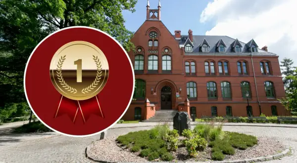 Złoty medal z cyfrą 1 na tle ceglanego budynku i zieleni.
