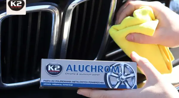 Ręce polerują chromowaną atrapę chłodnicy samochodu, używając pasty K2 Aluchrom. Efekt polerowania chromu jest widoczny.