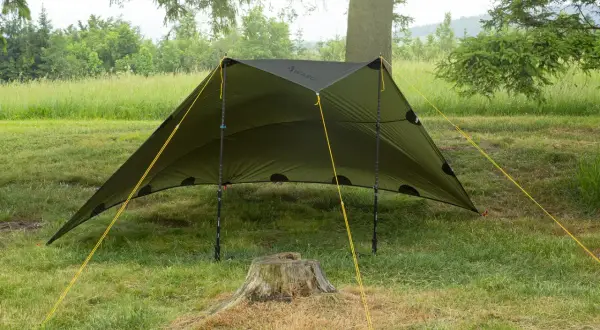 Zielony tarp rozstawiony na trawie, z żółtymi linkami. Pokazuje, jak zrobić namiot w prosty sposób, idealny na biwak.