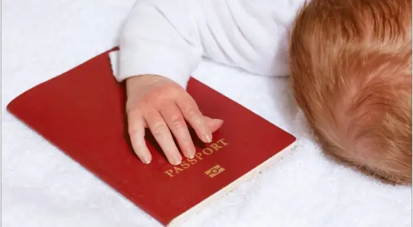 Dziecko z rudymi włosami śpi na białym ręczniku, jego dłoń spoczywa na czerwonym paszporcie. Czy do Hiszpanii potrzebny jest paszport?