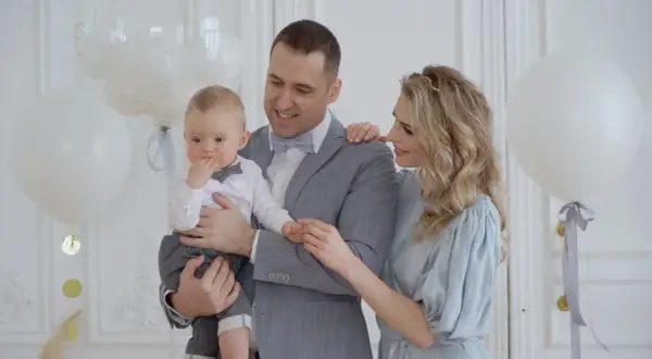 Uśmiechnięci rodzice trzymają synka w eleganckim stroju. Białe balony i złote konfetti tworzą atmosferę idealną na chrzciny dla chłopca.