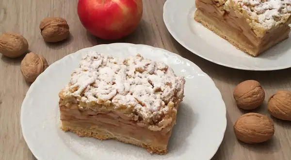 Szybka szarlotka z jabłkami ze słoika, posypana cukrem pudrem, obok orzechy i jabłko.