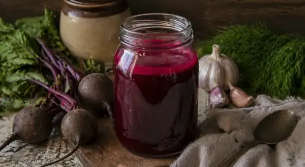 Słoik z kiszonym barszczem czerwonym, obok buraki, czosnek i koperek. Kiszony barszcz czerwony na co pomaga? Na odporność i trawienie!