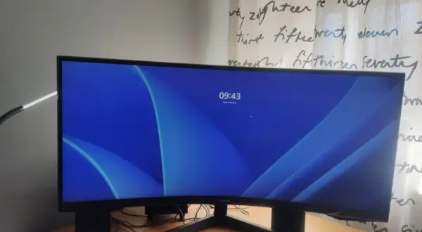 Zakrzywiony monitor 16:9 z niebieską tapetą i zegarem 09:43. Obok klawiatura i lampka.