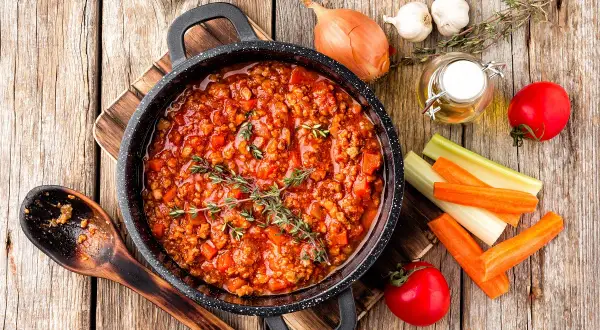 Pyszny sos bolognese w garnku, gotowy do podania z makaronem. Obok składniki: cebula, czosnek, marchew, seler, pomidory i oliwa. Idealny pomysł, co zrobić z sosem bolognese!