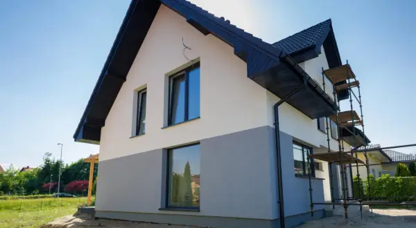 Nowy dom z dwukolorową strukturą elewacyjną, z szarym cokołem i białym tynkiem, w trakcie prac wykończeniowych.