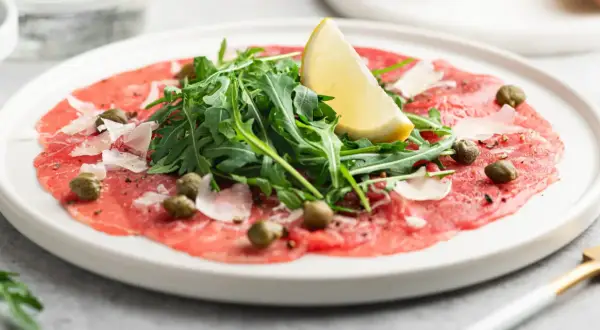 En tallrik med tunnskivad carpaccio di manzo, toppad med rucola, kapris, parmesan och en citronklyfta.