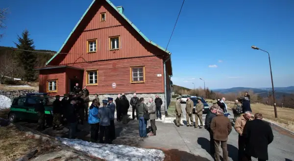 Grupa ludzi zgromadzona przed drewnianym budynkiem na przełęczy okraj. W tle góry i błękitne niebo.