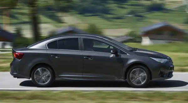 Ciemnoszara Toyota Avensis 2.0 D4D w ruchu, z rozmytym tłem górskiego krajobrazu.