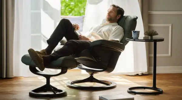 Man vilar i en bekväm Ekornes fåtölj med fotpall, njuter av ljuset från fönstret.