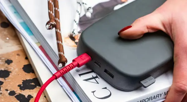 Ciemnoszary power bank 50000 mah na ile starcza? Podłączony czerwonym kablem, leży na stosie książek.