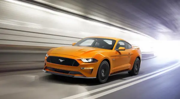 Un Mustang naranja acelera en un túnel, sus potentes sonidos de coches deportivos resonando.