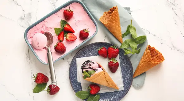 Erdbeer-Fruchteis selber machen: Ein Löffel Eis in einem Waffelhörnchen, garniert mit frischen Erdbeeren und Minze. Daneben eine Schale mit Eis und Erdbeeren.