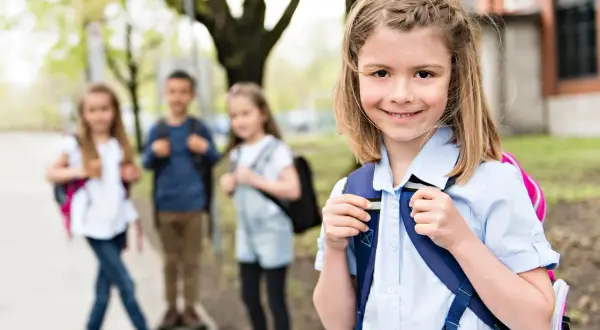 Uśmiechnięta dziewczynka z plecakiem, obok niej inni uczniowie. Czy można przenieść dziecko do innej szkoły?