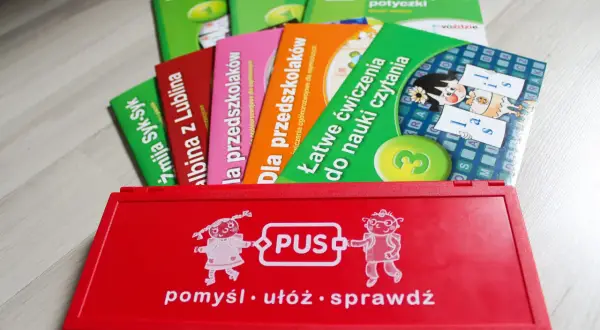 Czerwone pudełko z napisem "PUS" i rysunkami dzieci, obok kolorowe książeczki edukacyjne. Idealne do nauki i zabawy.
