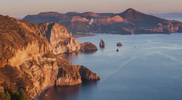 Scogliere dorate e mare blu intenso alle Isole Eolie, con piccole spiagge nascoste e faraglioni che emergono dall'acqua.