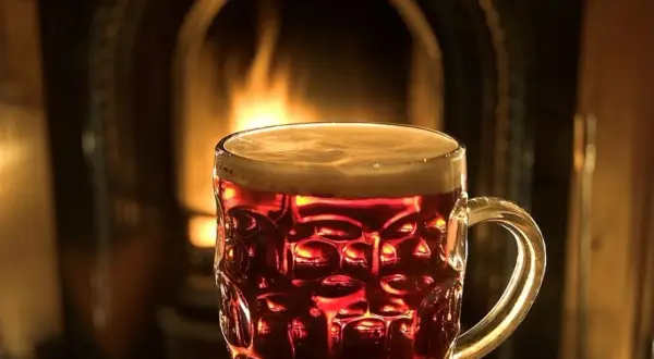 Bicchiere di birra ambrata con schiuma, davanti a un camino acceso. Un'atmosfera accogliente per gustare una birra calda.