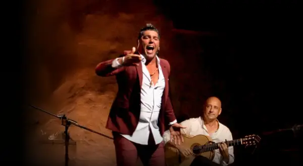 Un hombre con traje granate canta con pasión, su voz flamenco resuena mientras un guitarrista lo acompaña.