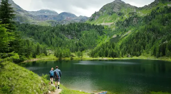 Para idąca ścieżką nad jeziorem, otoczonymi przez góry i lasy. W oddali widać szczyty, a na brzegu mała łódka. To piękne, górskie krajobrazy, gdzie można poczuć spokój i podziwiać przyrodę.