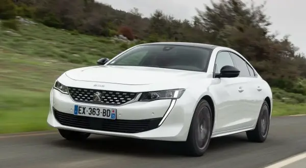 Biały Peugeot 508 jedzie po krętej drodze wśród zielonych wzgórz.