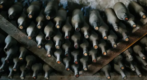 Des bouteilles de vin poussiéreuses, empilées sur des étagères en bois. La cave à vin semble avoir un problème de température, les bouchons sont secs.