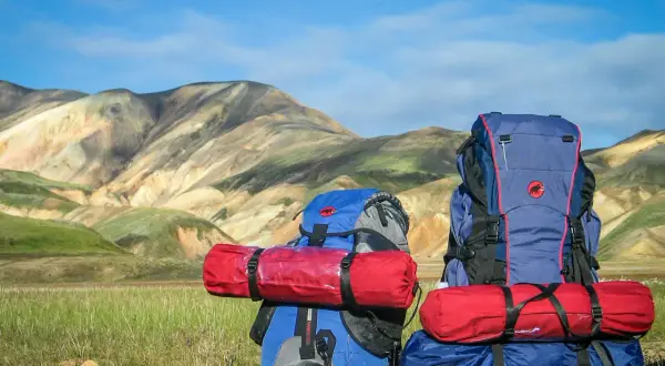 Dos mochilas de montaña listas para la aventura, con sacos de dormir rojos sujetos. El paisaje montañoso de fondo inspira a saber como llevar una mochila de montaña.
