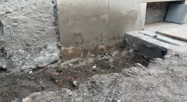 Prace przy fundamencie: odsłonięta ściana, ziemia i gruz. Widać fragmenty cegieł i beton. To etap prac, jak podlać fundamenty.